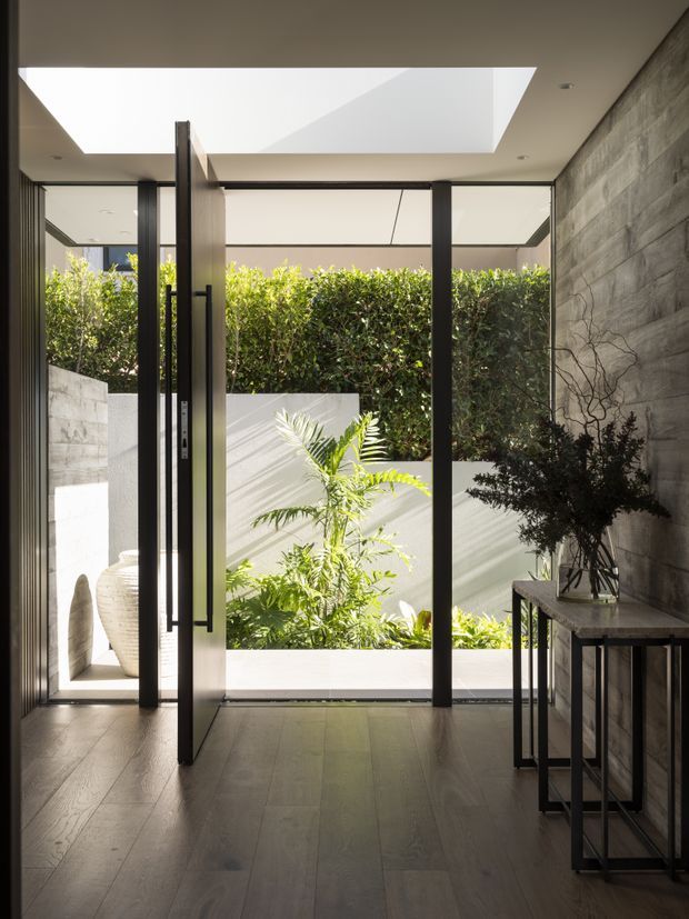 A refined modern entryway opening onto a small landscaped courtyard. The space features dark timber or wood-look flooring, a large pivot glass door with black metal framing, a concrete-textured feature wall, slim black console tables, and a broad skylight that brings in strong natural light. Tropical planting and a rendered privacy wall are visible beyond the glazing.