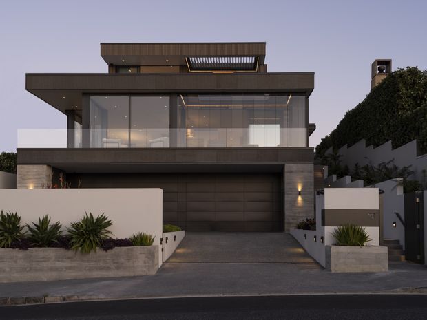 Contemporary two-storey urban residence viewed from the street at dusk. The facade features dark horizontal cladding, expansive floor-to-ceiling glazing, projecting flat roof forms, an integrated double garage, rendered boundary walls, and structured low-maintenance planting.