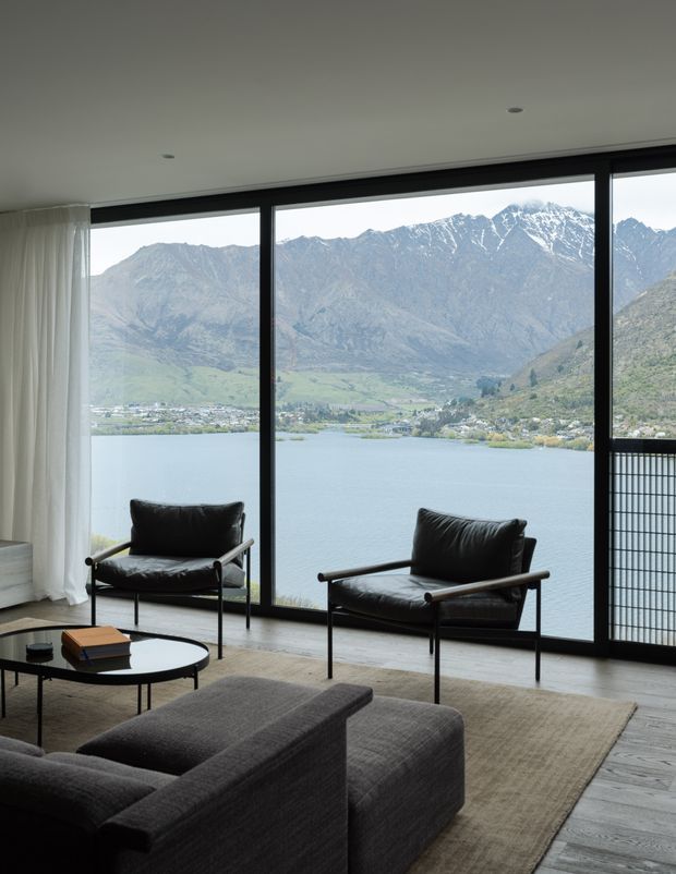 A refined living room with floor-to-ceiling black-framed glazing overlooking a lake and dramatic snow-dusted mountains. The room features two black leather lounge chairs, a dark charcoal sectional sofa, a low round coffee table, a large neutral rug, light timber flooring, sheer curtains, and a minimalist recessed ceiling.