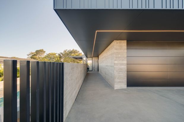 A modern residential exterior showing a covered driveway or carport passage beside a garage. The space features a broad cantilevered dark soffit with a linear recessed LED detail, smooth light concrete or stone walls, a large dark horizontal-panel garage door, and a tall vertical dark metal privacy fence. Minimal landscaping and trees are visible beyond the fence.