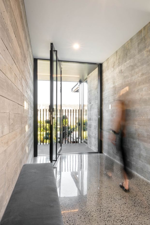 A narrow, minimalist interior passage or entry hall with exposed grey concrete block walls, a polished speckled concrete floor, and black-framed glazed doors opening to a balcony or exterior walkway. A built-in grey upholstered bench lines the left wall, while bright daylight and vertical exterior screening are visible beyond the doors. A blurred person is walking through the space.