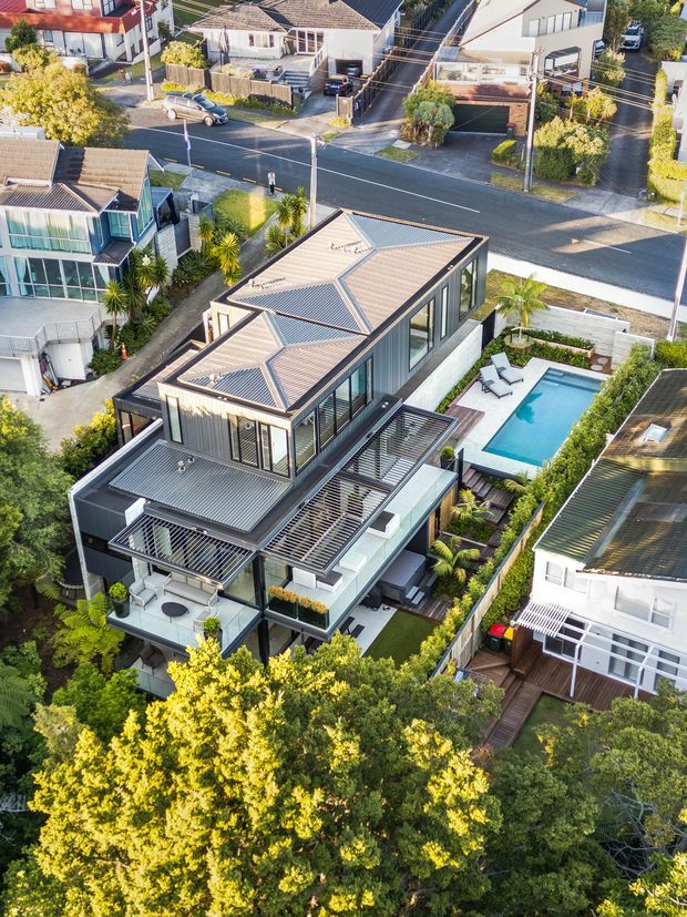 Aerial view of a contemporary multi-level residential house in a leafy suburban neighbourhood. The home has a complex pitched metal roof, extensive floor-to-ceiling glazing, dark exterior framing, multiple balconies and terraces, landscaped gardens, and a rectangular swimming pool beside a timber deck. Surrounding detached houses and streets are visible.