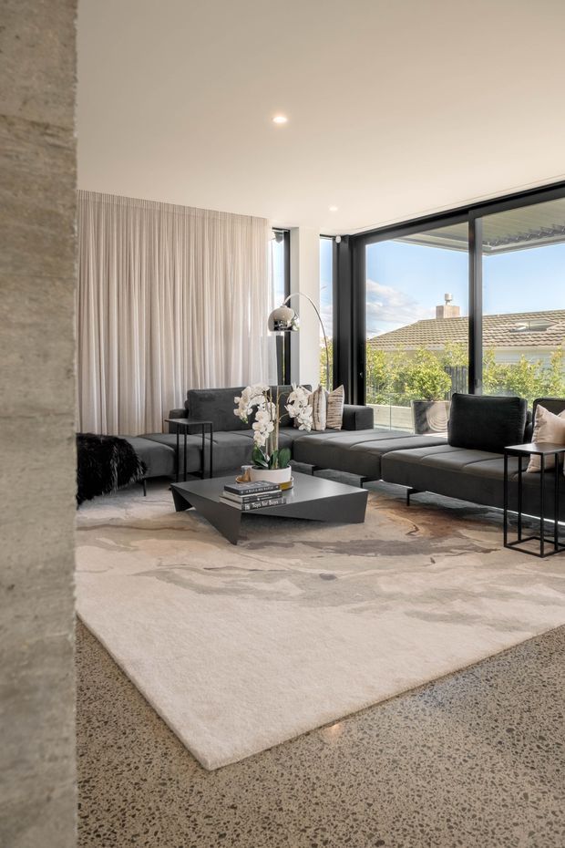Contemporary living room with a large dark modular sectional sofa, sculptural black coffee table, slim side tables, floor-to-ceiling black-framed glazing, sheer full-height curtains, and a softly patterned neutral rug. The room has a polished concrete floor, a high plain white ceiling with recessed downlights, and a textured concrete or stone wall at the left. Suburban rooftops and trees are visible beyond the glazing.