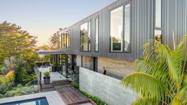 Exterior view of a contemporary multi-level residence on a landscaped, sloping site. The building features dark vertical metal cladding, extensive floor-to-ceiling glazing, exposed concrete retaining walls, timber decking and a partially visible swimming pool, with lush tropical planting surrounding the outdoor areas.