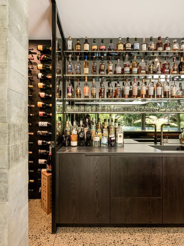 A sophisticated built-in home bar with extensive glass shelving displaying liquor bottles and glassware. Dark timber cabinetry supports a stone or stainless-look benchtop with an integrated sink and tap. A dedicated wine rack is visible to the left, while a large window behind the bar brings in greenery and daylight. The space has a refined, contemporary feel with terrazzo-style flooring and a pale stone-clad wall.
