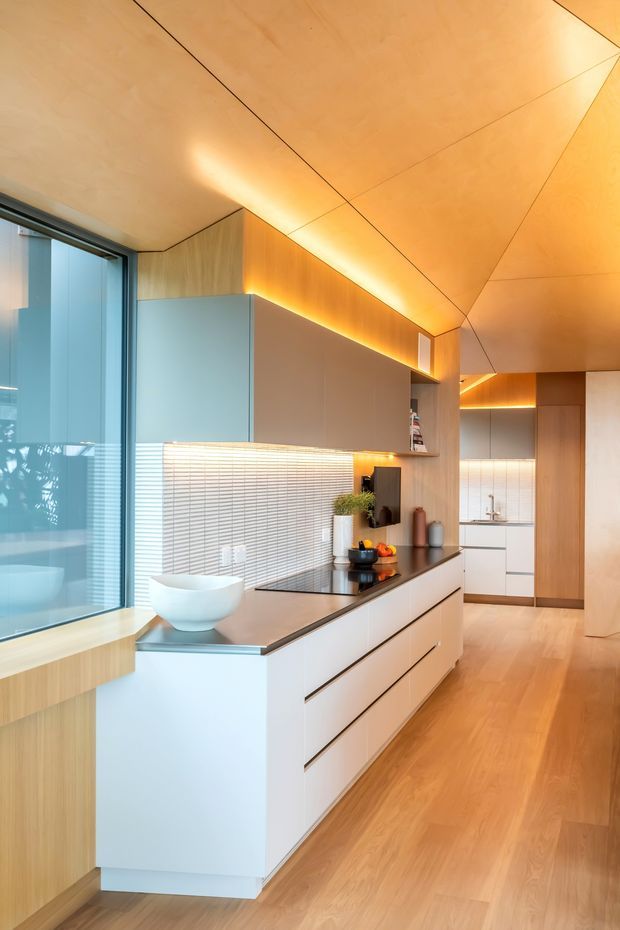 A refined contemporary kitchen with a long white island and integrated drawers, dark benchtop, pale timber cabinetry, and a white horizontal tile splashback. Warm concealed LED lighting highlights the timber-lined ceiling and upper joinery. Full-height glazing brings daylight into the compact, elongated space, with timber flooring continuing throughout.