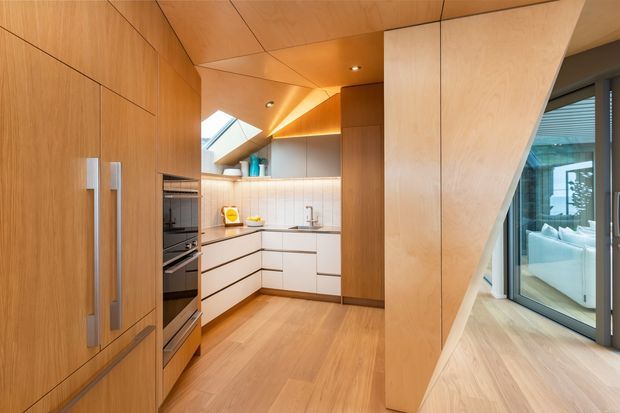Compact contemporary kitchen with warm timber-lined cabinetry, walls, ceiling and flooring. The kitchen features white lower cabinetry, integrated stainless-steel appliances, a pale tiled splashback, timber benchtops or trim, and a skylight. A sculptural angled timber partition frames glazing and a glimpse of an adjacent furnished living area.