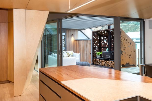 View from a contemporary kitchen toward a glazed living area and courtyard-like outdoor room. The image features warm timber cabinetry and ceiling finishes, a large timber kitchen island with a pale stone benchtop, dark-framed glazing, and a sculptural timber screen surrounding a fireplace and stacked firewood.