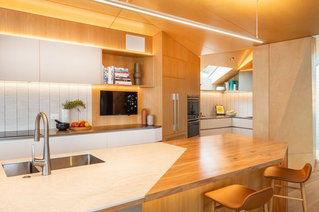 Warm contemporary kitchen with extensive timber cabinetry and ceiling panelling, a pale stone island with integrated sink and gooseneck tap, timber breakfast bar, built-in appliances, open shelving, and a compact dining or living area visible beyond. Skylights and linear lighting provide a bright, inviting atmosphere.