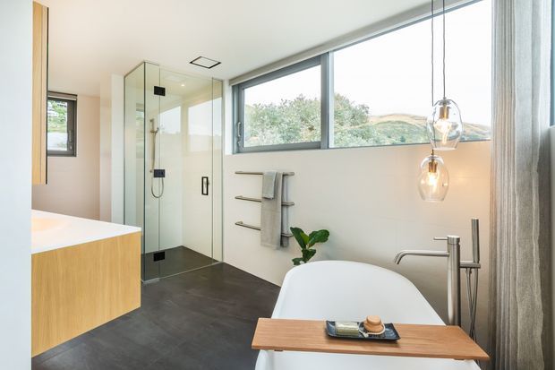 A bright contemporary bathroom with a freestanding white bathtub, wall-mounted timber bath tray, frameless glass walk-in shower, wall-hung towel rails, floating timber vanity, and dark flooring. Large horizontal windows bring in natural light and overlook greenery, while pendant lights hang beside the bath.