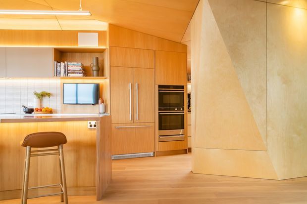 Warm, compact contemporary kitchen interior with extensive light timber cabinetry and matching timber ceiling and flooring. The kitchen includes a peninsula with seating, integrated refrigerator and double oven, open shelving, white tiled splashback, and a sculptural pale textured partition wall. A linear ceiling light and concealed warm lighting create a cohesive wood-lined fit-out.