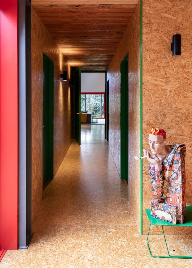 A narrow interior hallway lined with exposed oriented strand board (OSB) walls, ceiling, and flooring. Deep green door frames and trim contrast with vivid red accents, while a bright window at the far end provides natural light. A colourful sculptural figure sits on a green-framed display stand in the foreground.