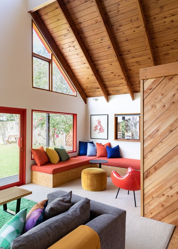 A bright, colourful living room with a dramatic pitched, timber-lined ceiling and exposed timber rafters. The room features a built-in red window seat with multicoloured cushions, a grey sofa, mustard ottoman, red sculptural chair, timber-framed glazing and a large diagonal timber-clad partition or door. The space opens visually to a landscaped garden outside.
