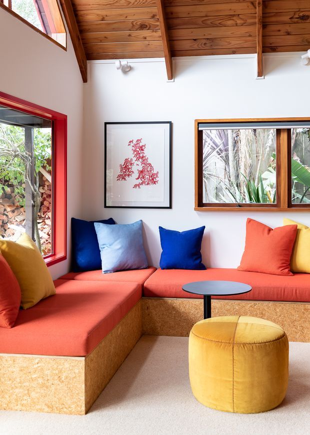 A compact, colourful living room with built-in L-shaped seating upholstered in coral-red fabric and faced with exposed wood-fibre board. Bright blue, light blue, orange, red, and yellow cushions create a playful palette. The room features a warm timber-lined pitched ceiling with exposed rafters, white walls, red-painted window and door frames, timber-framed glazing, and a small round black side table beside a yellow cylindrical ottoman. Lush planting is visible outside the windows.