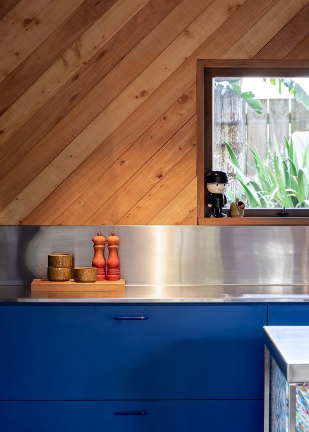 Close-up view of a compact kitchen featuring vivid blue fixed cabinetry with dark handles, a brushed stainless-steel splashback, and warm diagonal timber wall panelling. A timber-framed window brings in natural light and overlooks greenery, while a small timber display board with orange-red salt and pepper mills sits on the benchtop. The image has a warm, playful, richly contrasting material palette.