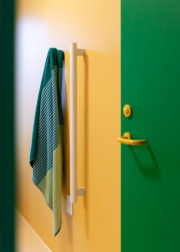 Close-up interior view of a brightly coloured bathroom or utility-space wall beside a green door. A teal and striped towel hangs from a tall white wall-mounted towel rail, with a yellow door handle and lock providing bold contrasting accents against the warm yellow wall.