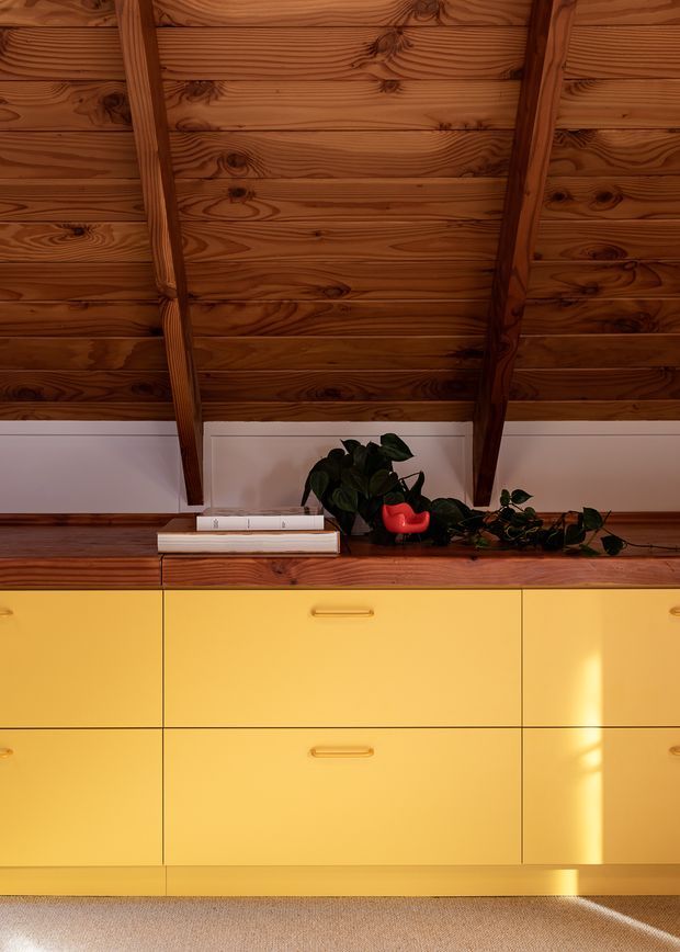 Close-up interior view of a warm, richly grained timber ceiling with exposed rafters above a built-in yellow cabinet or sideboard. The timber countertop holds books and potted plants, while sunlight creates bright highlights across the cabinetry and carpeted floor.