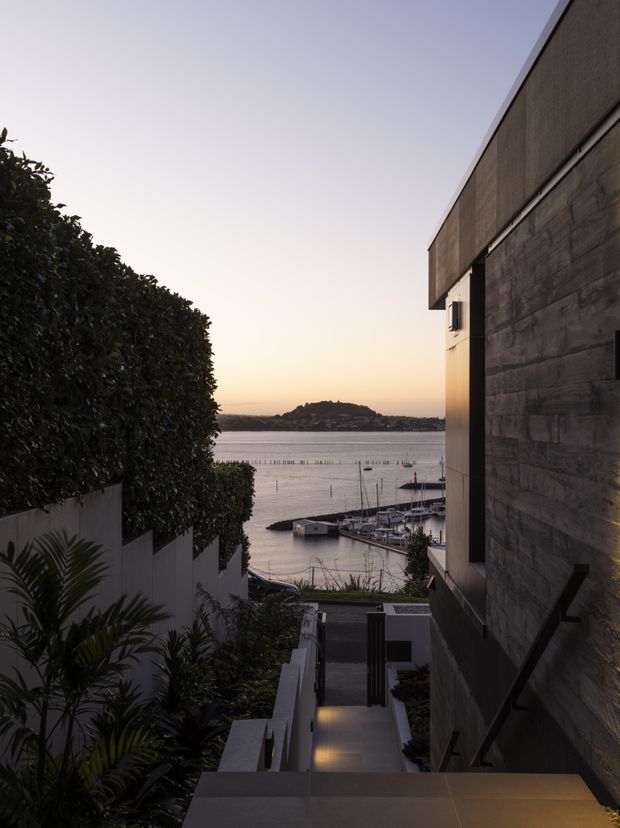 A dusk exterior view down a landscaped stairway beside a contemporary waterfront residence. The building has a dark textured concrete or stone-like wall, projecting upper volume, and minimalist detailing. Beyond the property is a marina and calm inlet with a low, vegetated headland on the horizon. Dense hedging and tropical planting line the stairs, with warm recessed step lighting creating a refined, tranquil atmosphere.
