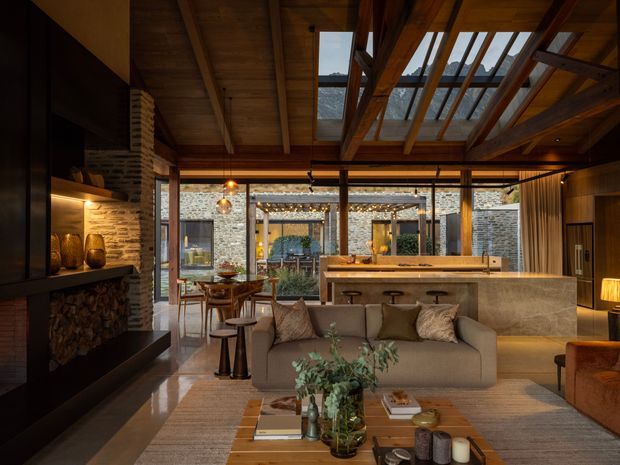 A warm, sophisticated open-plan living space combining a lounge, dining area and kitchen. The room features extensive exposed timber structure, a dramatic glazed roof section, dark timber cabinetry and wall cladding, a large stone kitchen island, polished stone flooring, a built-in fireplace with stacked firewood, and full-height glazing opening toward a landscaped stone courtyard. Neutral upholstered seating and layered ambient lighting create a cozy, luxurious atmosphere.