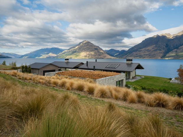 A low, modern single-storey house with dark metal pitched roofs sits on a grassy lakeside site. A textured grey masonry courtyard or retaining wall is visible in front, surrounded by golden tussock grasses and landscaped lawn. The home overlooks a vivid blue lake framed by steep rugged mountains beneath a partly cloudy sky.