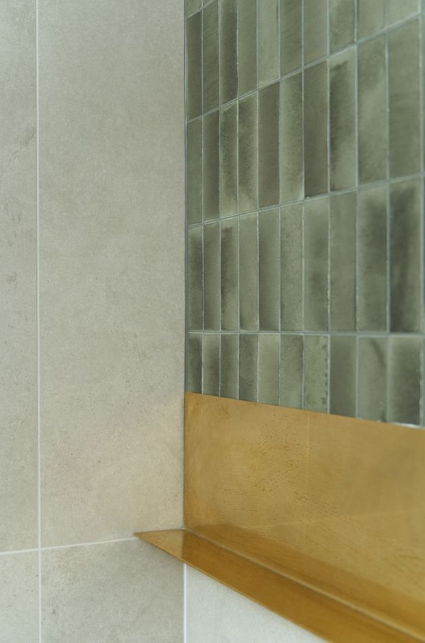 Close-up detail of a bathroom wall junction featuring large pale grey stone-look tiles, a vertical section of smaller glossy grey-green rectangular tiles, and a warm honey-toned timber ledge or trim running horizontally along the base of the tiled feature wall.
