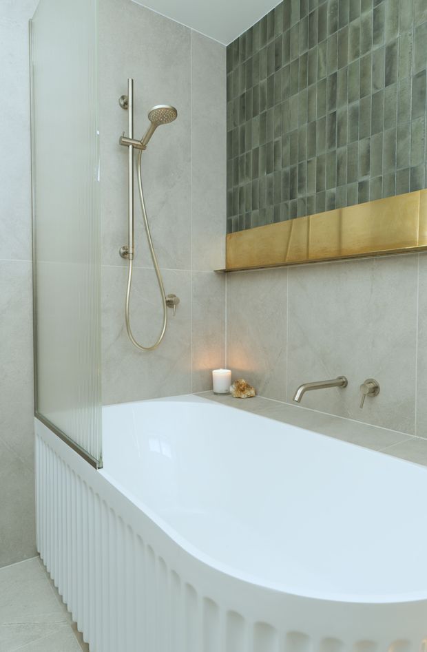 A refined bathroom featuring a white integrated bathtub with a fluted front panel, a wall-mounted brushed brass shower and bath tapware, pale grey stone-look wall tiles, and a dark green-grey tiled feature wall. A brass-toned recessed shelf runs across the feature wall, with subtle candle styling adding warmth.