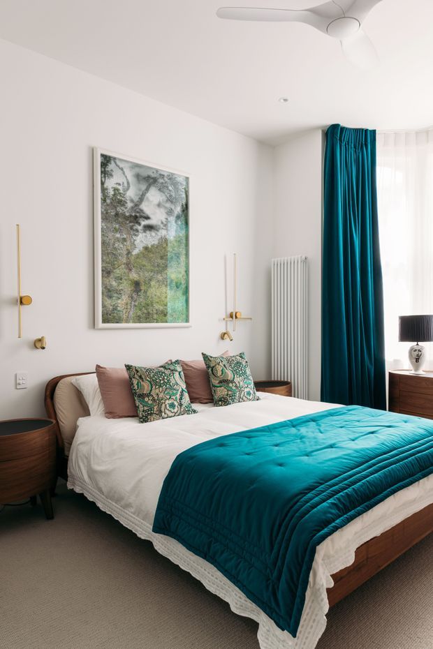 A bright, refined bedroom with a large upholstered bed dressed in white bedding, patterned decorative cushions, and a deep teal quilt. White walls and ceiling create an airy backdrop, complemented by matching teal floor-to-ceiling curtains, a tall white radiator, warm brass wall sconces, dark wood bedside tables, and a framed botanical artwork. A contemporary ceiling fan and softly filtered window light complete the room.