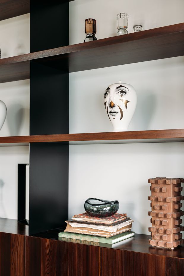 Close-up interior view of a custom built-in display shelving unit. Dark timber shelves and richly grained timber cabinetry are framed by matte black vertical supports and a white painted backing. The shelves display decorative glassware, a sculptural face vase, a dark glass bowl, books, and a textured wooden vessel, creating a curated, gallery-like composition.