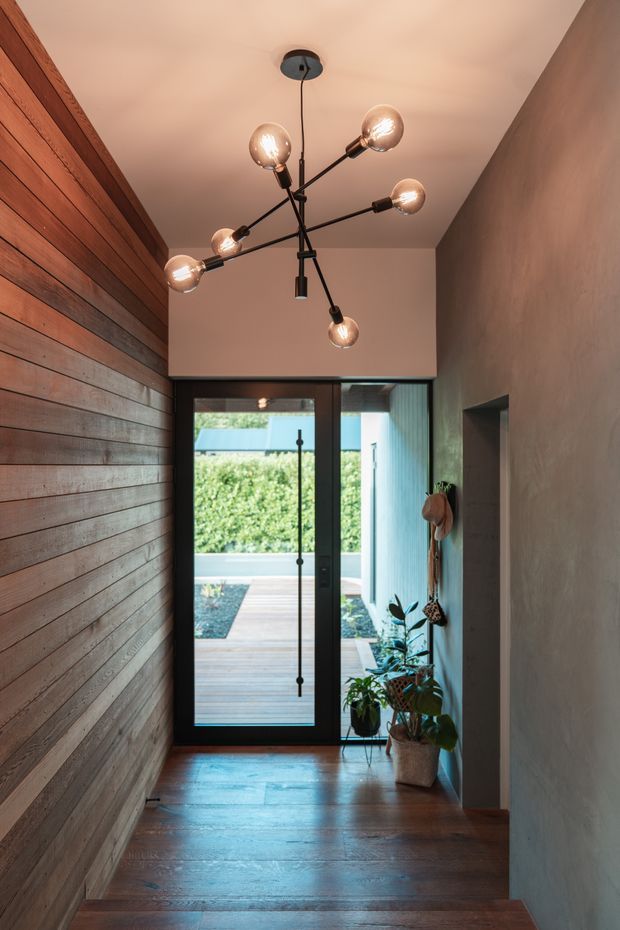 A narrow, warmly lit residential entry hallway with dark timber flooring, a horizontal timber feature wall on the left, smooth plaster walls, and a striking multi-arm globe pendant light. A full-height glazed black-framed door at the end opens to a small timber deck and landscaped courtyard. Potted plants and wall-mounted accessories add natural detail.