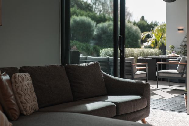 A contemporary living room viewed from beside a dark upholstered sofa, opening through large black-framed sliding glass doors to a furnished outdoor terrace. The interior has neutral walls, a textured decorative cushion, and a pale rug, while the terrace includes outdoor lounge and dining furniture surrounded by dense greenery.