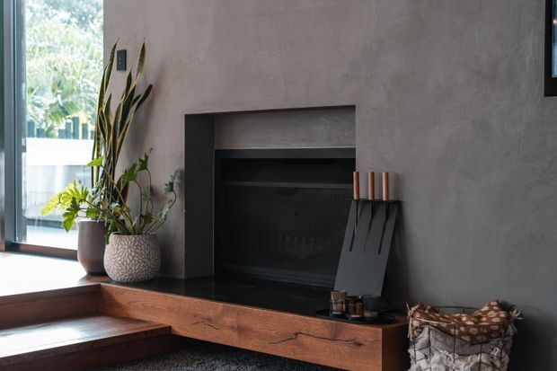 A contemporary living room fireplace set into a textured grey plaster wall, with a dark recessed firebox, black metal fireplace tools, and a substantial warm timber hearth bench. Potted indoor plants sit beside the hearth, while timber steps and a large window bring natural light into the space.