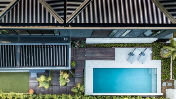 Aerial top-down view of a contemporary residential property showing a rectangular swimming pool, pale paved pool surround, dark timber decking, landscaped planting, and a modern building with strong linear roof and facade elements. The composition emphasizes the outdoor entertaining and pool area.