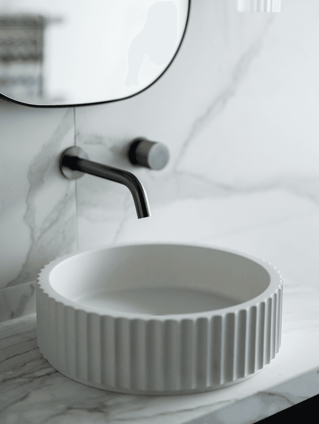 Close-up of a refined bathroom vanity featuring a round fluted white vessel basin on a veined white marble countertop. A minimalist black wall-mounted tap and matching circular control sit against a white marble slab backsplash, with a rounded-edge mirror above.