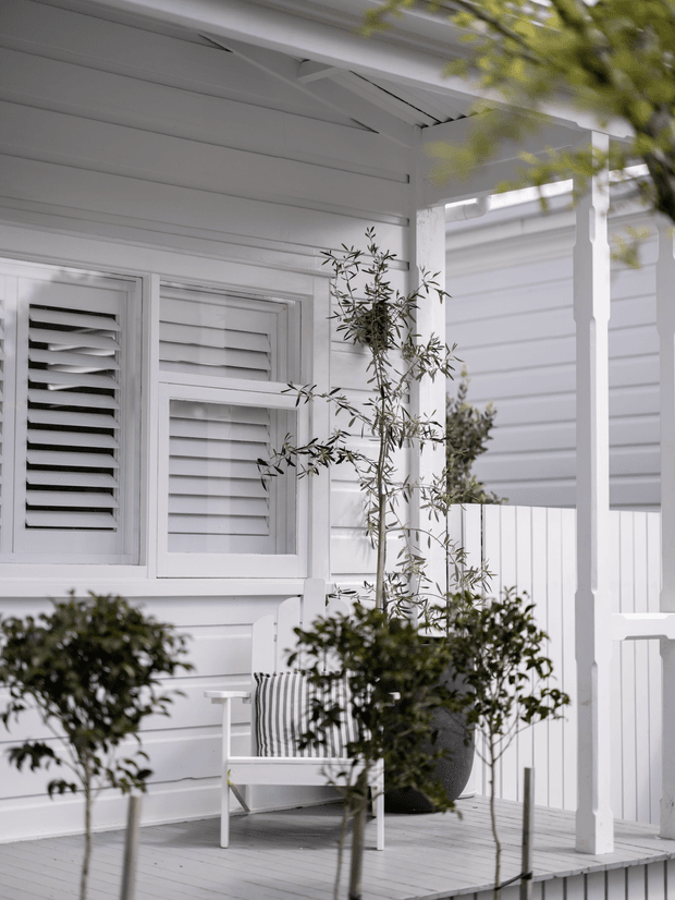 A bright white-painted residential veranda with horizontal weatherboard cladding, louvred plantation-shutter windows, a covered porch roof, square posts and balustrades, and a small white outdoor chair with striped cushion. Potted and planted greenery softens the crisp coastal-style exterior.