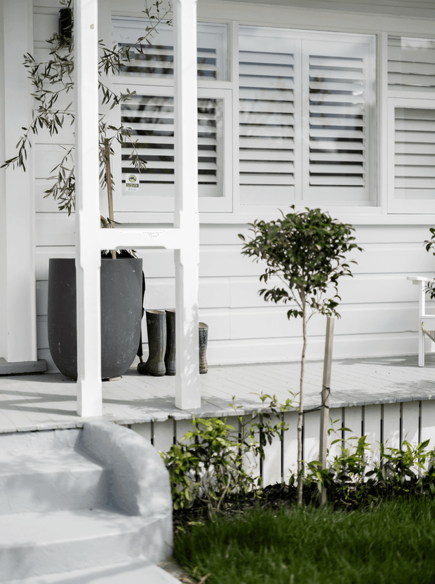 A bright white-painted porch and entry deck with horizontal weatherboard cladding, shuttered windows, simple square posts, white decking and steps, and neatly planted shrubs. Large dark planters and boots sit near the entry, while a small tree and garden border frame the foreground.