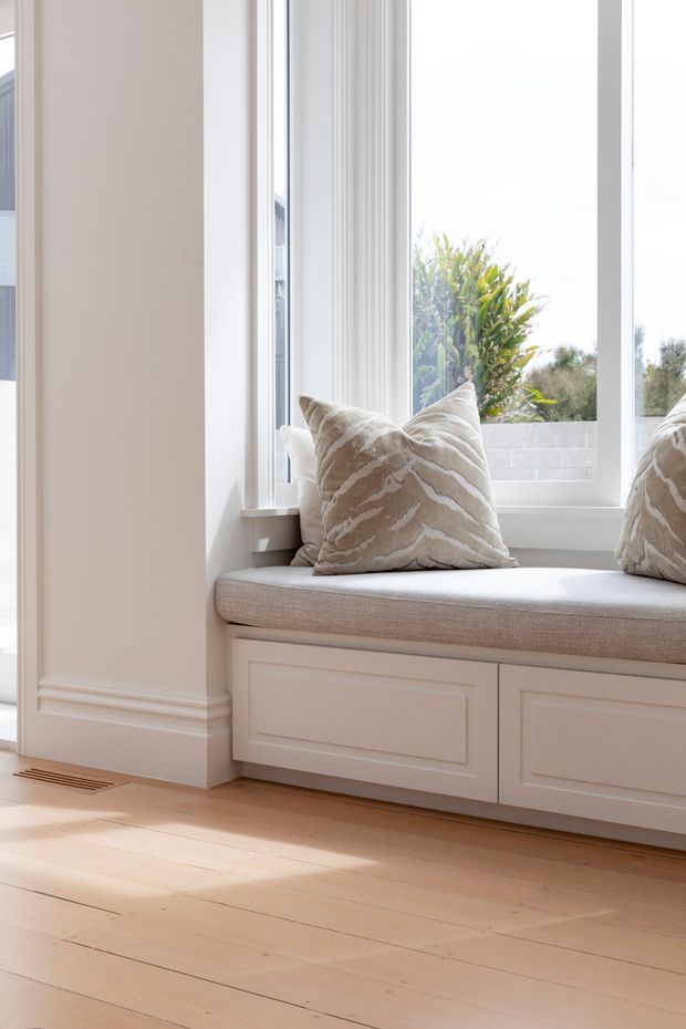 Bright, refined interior detail showing a built-in window seat beneath large white-framed windows. The seat has pale upholstered cushioning and neutral patterned pillows, with white paneled cabinetry below. Light timber floorboards, warm off-white walls, and traditional decorative skirting create an airy, elegant atmosphere. Garden planting and a rendered exterior wall are visible outside the windows.