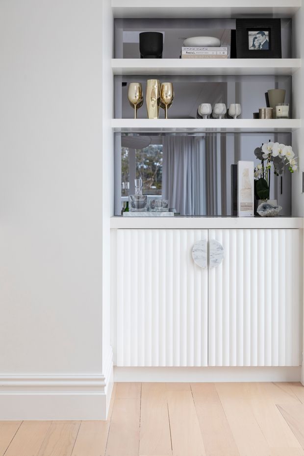 A refined built-in display and storage unit set into a pale wall. Open white shelves hold glassware, books, decorative objects and framed photography, while a mirrored back reflects a bright interior with curtains. The lower section features white fluted cabinet doors with round stone-like pulls, above a light timber floor. The overall composition is bright, restrained and contemporary with subtle classic detailing.