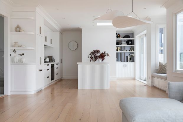 Bright open-plan interior combining a white kitchen with integrated cabinetry, a central fluted island, built-in shelving, and a light-filled lounge area. Pale timber flooring, white panelled joinery, decorative cornices, and sculptural pendant lights create a refined, calm, contemporary-classic atmosphere.