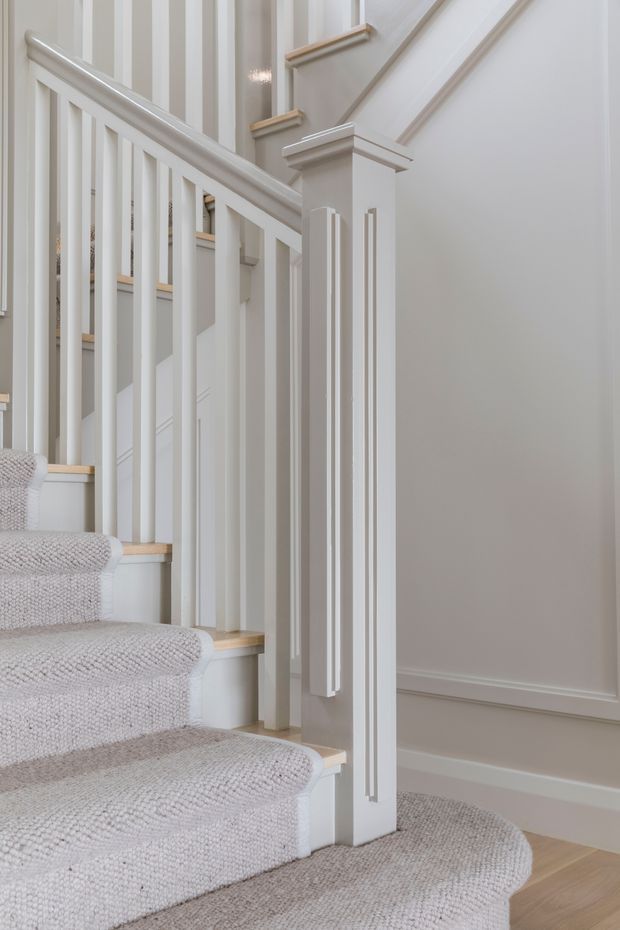 Close-up interior view of a refined staircase with pale textured carpeted treads, white painted balustrades, turned-style decorative newel detailing, and traditional wall panelling with moulded trim. The space is bright, neutral, and carefully finished, with timber flooring visible at the base.