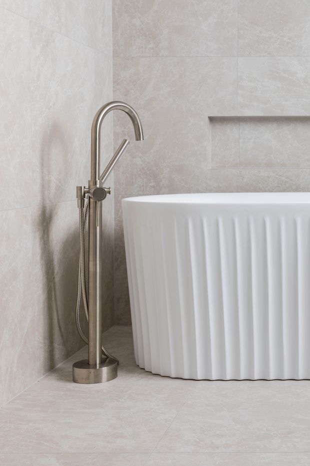 Close-up of a refined bathroom featuring a freestanding white bathtub with vertical fluted detailing, a brushed metallic floor-mounted bath mixer with hand shower, large pale stone-look wall and floor tiles, and a recessed wall niche.