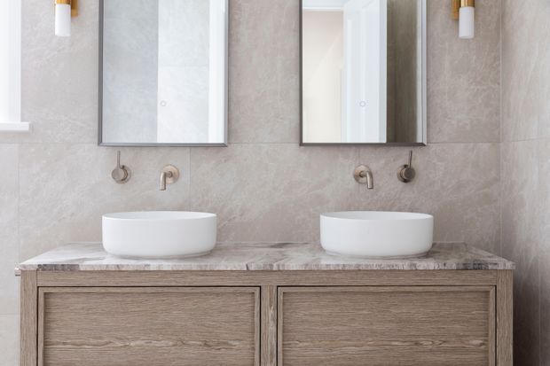 A refined double-vanity bathroom with two white vessel basins set on a grey-beige stone benchtop, paired with wall-mounted brushed metal taps. The vanity has twin timber-look drawer fronts, while large pale stone-look wall tiles and rectangular mirrors create a calm, cohesive finish. Vertical wall sconces with warm brass-toned details flank the mirrors.