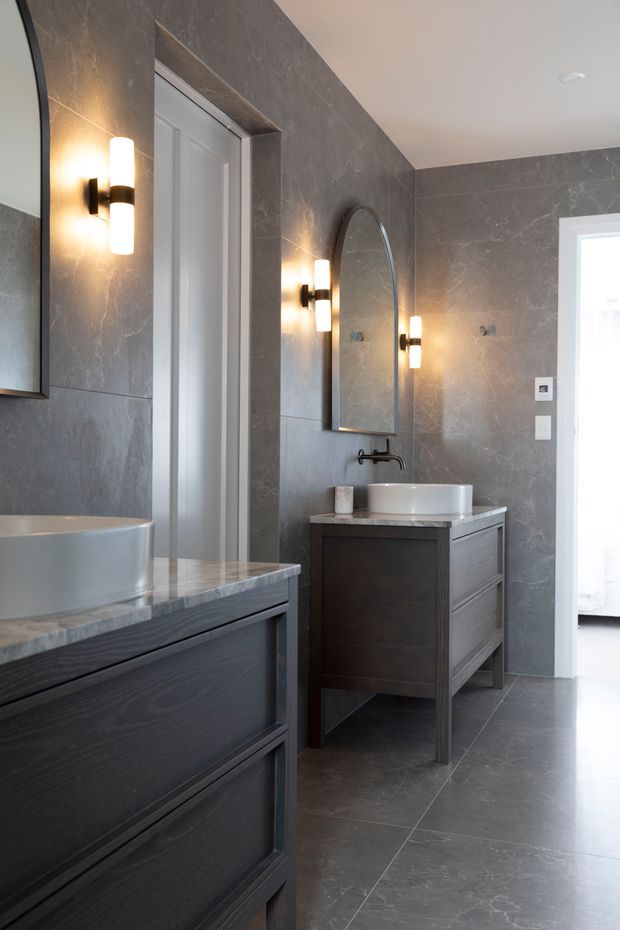 A refined contemporary bathroom with dark grey stone-look wall and floor tiles, a dark timber vanity with drawers, a white vessel basin and a matching freestanding bathtub partially visible in the foreground. Arched mirrors and cylindrical wall sconces add a classic accent, while a doorway opens toward a bright adjoining room.