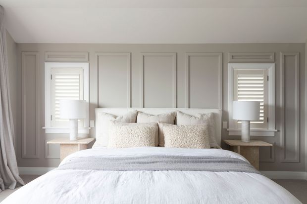 A bright, symmetrical bedroom with a neatly made bed centered against a grey paneled feature wall. The room includes white bedding, layered neutral pillows, matching bedside tables and lamps, plantation shutters, and full-height curtains. The overall design is calm, refined, and softly neutral.