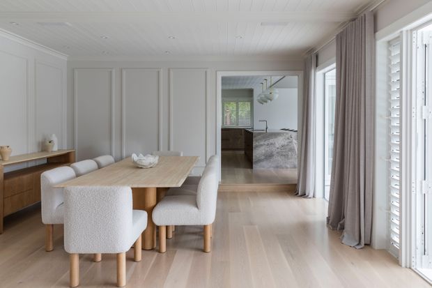 Bright, refined dining area with a light timber dining table and upholstered chairs, set against a white paneled wall and pale timber flooring. Full-height glazed doors with plantation shutters and soft taupe curtains bring in abundant natural light. A kitchen with a stone island is visible beyond the opening.