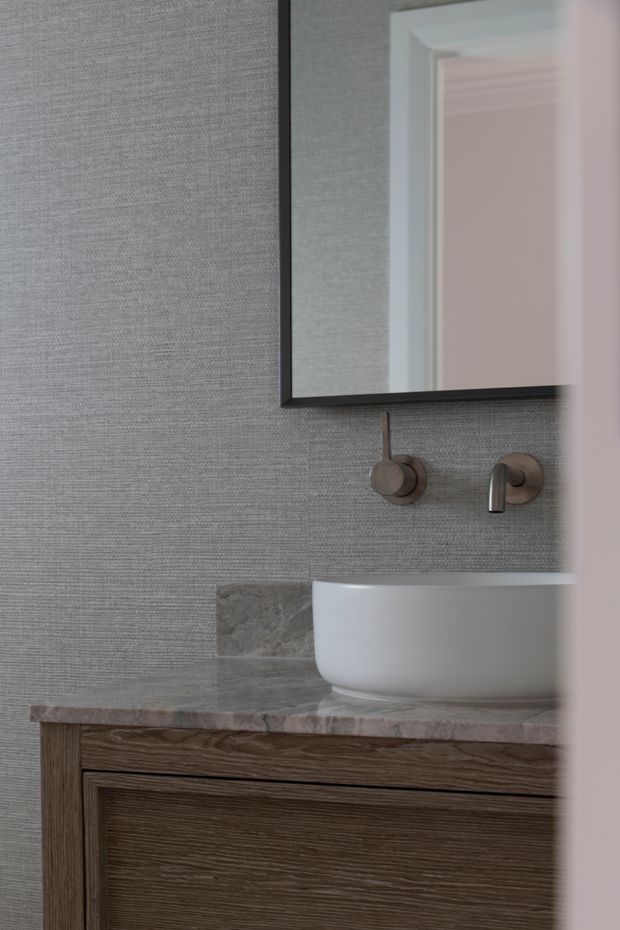 Close-up view of a refined bathroom vanity. A white vessel basin sits on a grey-veined stone benchtop above dark timber-grain cabinetry, with wall-mounted brushed metal tapware and a black-framed rectangular mirror. The walls have a textured grey finish, and a partially visible doorway appears at right.
