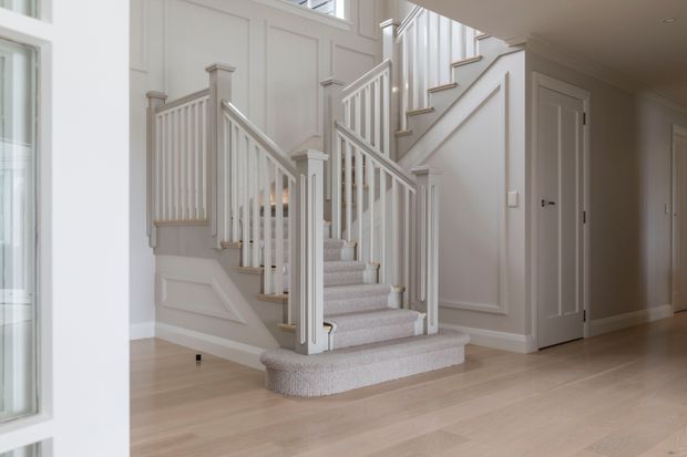 Bright, refined residential stair hall featuring a split staircase with white painted turned posts, balusters, and panelled wall detailing. The stairs are carpeted in a light neutral tone, while the surrounding floor is pale timber. White doors, trim, and abundant glazing create an airy, traditional interior.