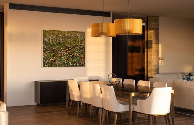 Warm contemporary open-plan dining and living area with a long dark timber dining table, upholstered dining chairs, a linear sideboard, sculptural pendant lights, timber flooring, and a neutral sofa. A textured dark feature wall and stone-clad column add contrast, while artwork and late-afternoon light create an inviting atmosphere.