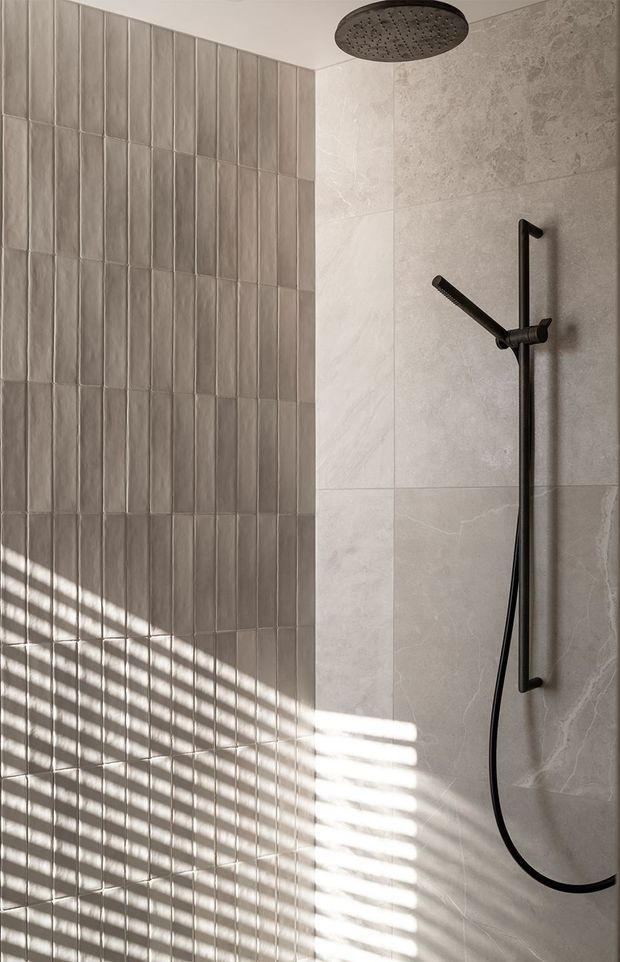 Close-up of a contemporary walk-in shower, featuring large pale stone-look wall tiles and a contrasting vertical field of narrow textured rectangular tiles. A matte black rainfall showerhead, handheld shower rail and hose are mounted on the wall. Strong sunlight casts linear shadows across the tiled surface.