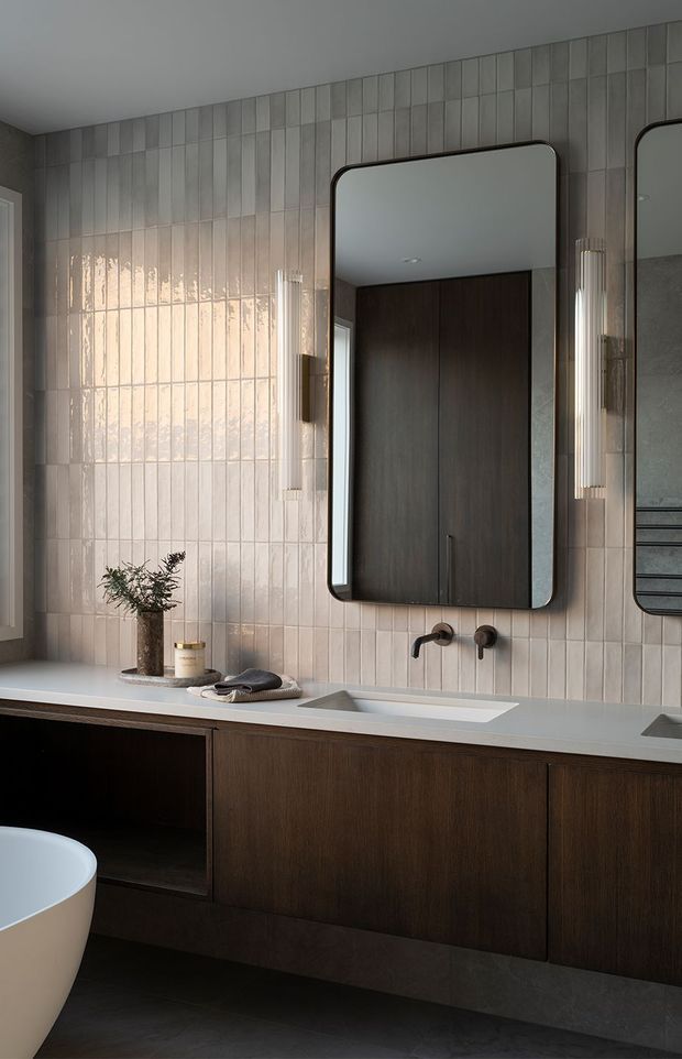 A refined contemporary bathroom featuring a long double vanity with dark timber cabinetry, pale stone benchtop, integrated rectangular basins, wall-mounted matte-black tapware, and rounded-rectangle mirrors. Glossy vertically arranged light grey-beige wall tiles create a reflective backdrop, complemented by slim ribbed wall sconces and a partially visible freestanding bathtub.