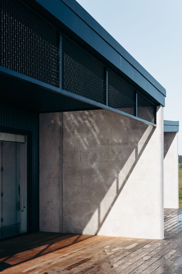 A close-up exterior architectural view of a contemporary building, featuring a dark metal-clad upper level with fine perforated or mesh screening, a raw textured concrete wall, and a timber deck. Strong angular sunlight creates geometric shadows across the wall and decking.